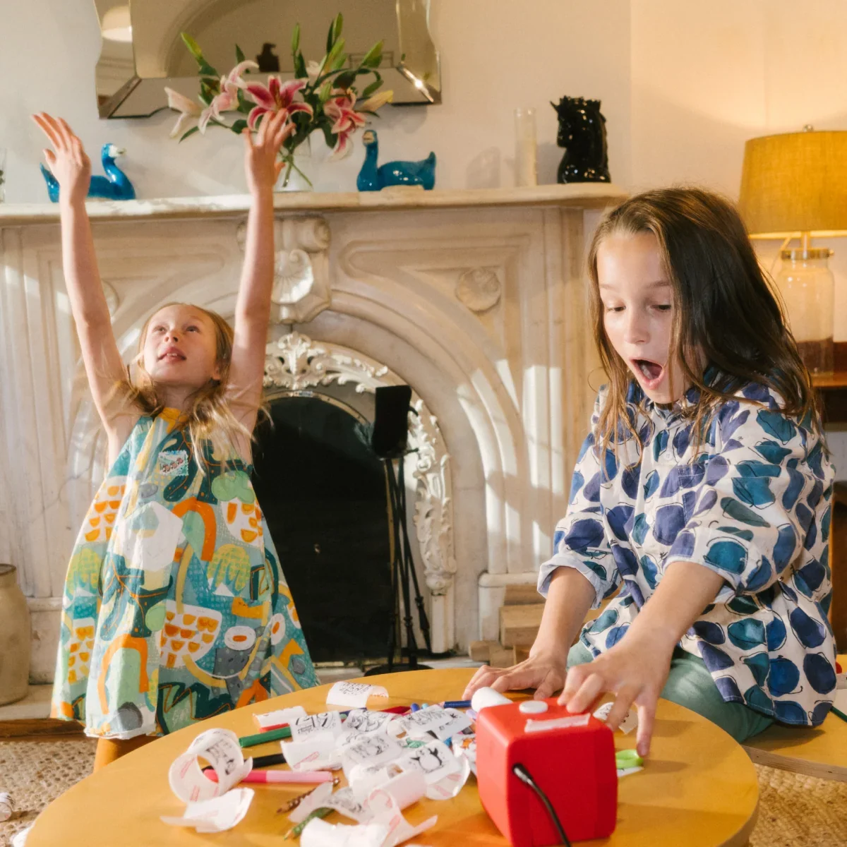 Two young girls are playing with Stickerbox in a cozy living room. The girl on the left is wearing a colorful patterned dress and has her arms raised in excitement, while the girl on the right, wearing a blue and white patterned shirt, is looking at Stickerbox.