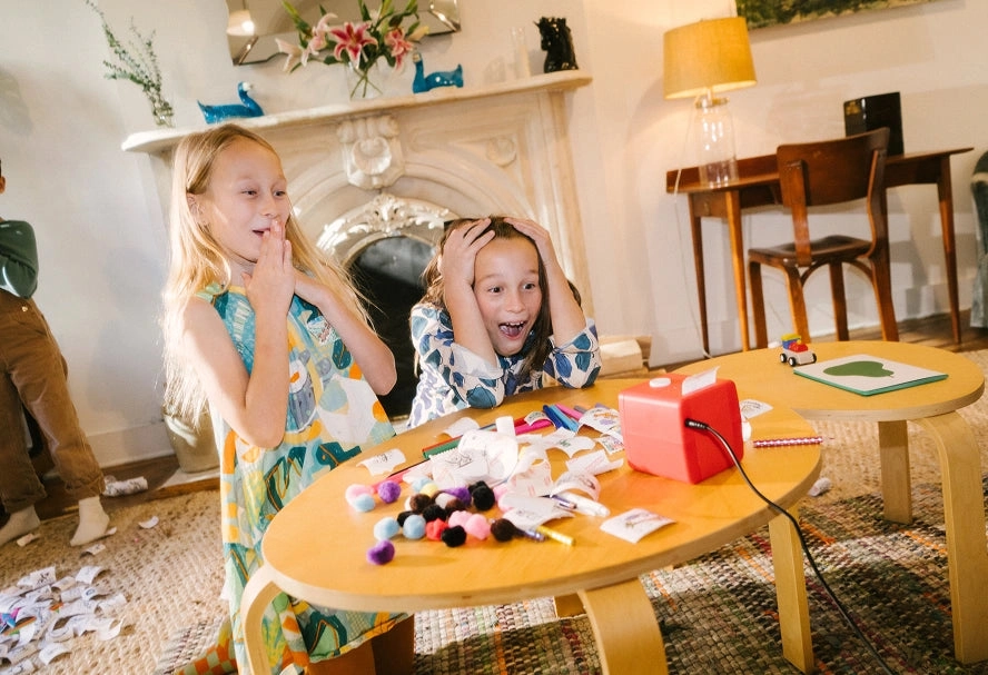 Two girls gasp with their hands on their faces at a table covered in stickers, pencils and a red Stickerbox