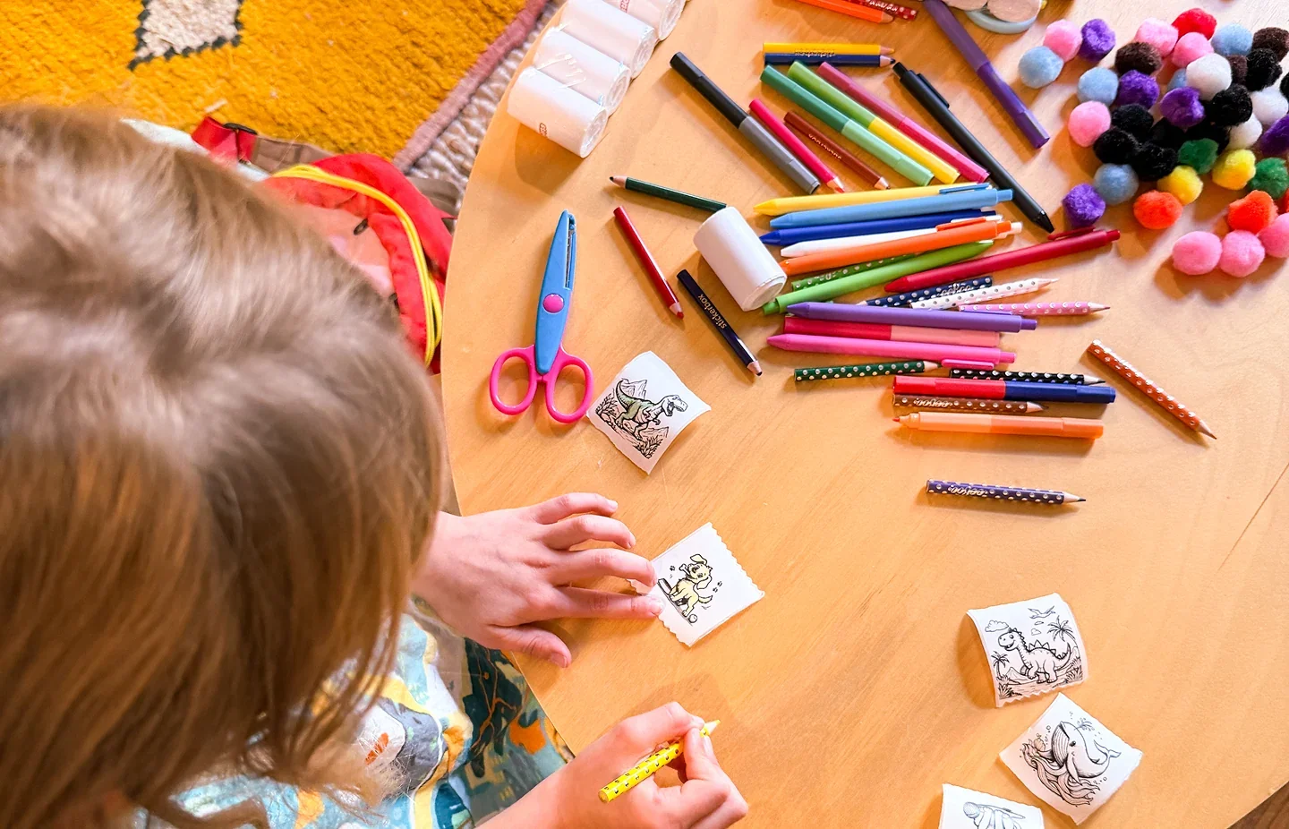 Children at a wooden table coloring black-and-white stickers with pencils and scissors