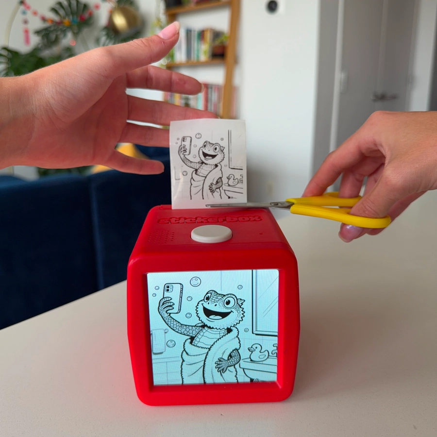 Two hands at a Stickerbox: one pulls the freshly printed sticker, the other holds yellow kid-safe scissors