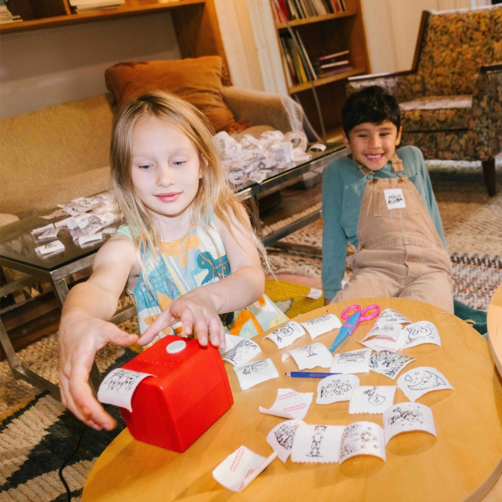 Two children at a round table sharing a red Stickerbox and a pile of freshly printed stickers