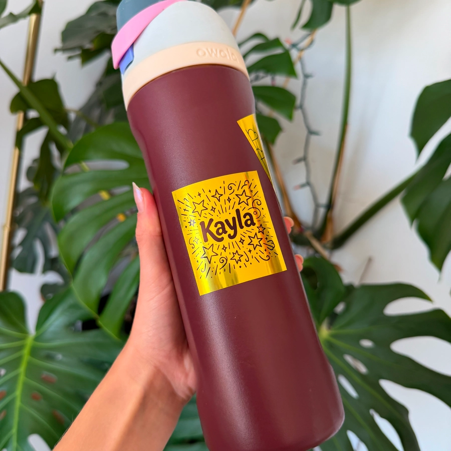 A gold holographic name sticker on a dark red water bottle held in front of a plant